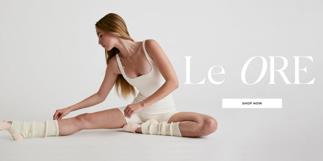 Model wearing a white short catsuit from Le ore with legwarmers and ballet shoes on a grey background with the words Le Ore: Shop Now
