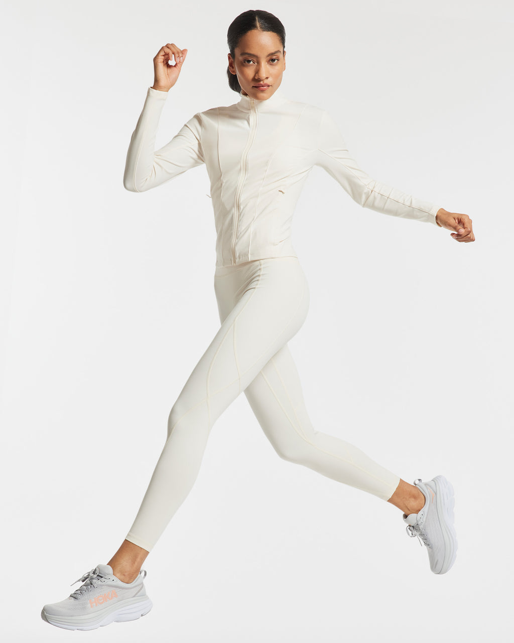 Woman wearing all white activewear and Hoka sneakers running in front of a white background.